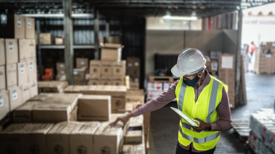 Warehouse worker wearing face mask and protective workwear checking products using a tablet