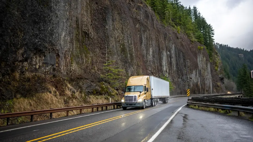 Yellow semi truck transporting cargo on a wet road with a rock cliff wall on the side