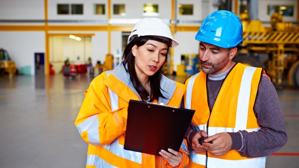 two warehouse workers talking together over a clipboard while standing inside of a large warehouse