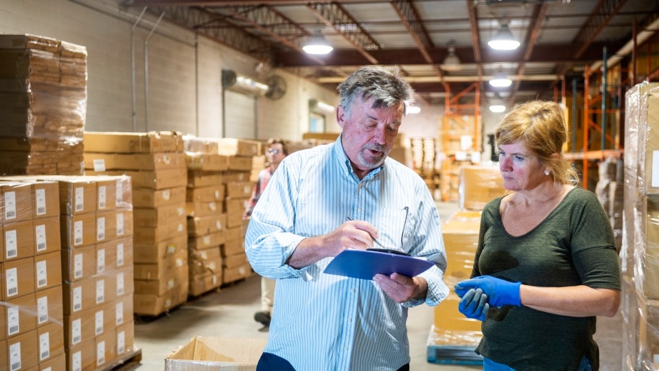 A warehouse manager and an floor worker having a discussion in the warehouse as another employee walks by holding a box